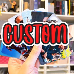 May include: A hand holding a stack of custom photo cards. The cards feature various images and are partially obscured by a large, red and black "CUSTOM" text overlay. The background includes a bookshelf with figurines and books.