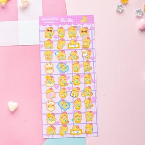 Op de afbeelding: Een vel stickers met gele cartoon eend-personages in verschillende poses, met accessoires zoals zonnebrillen en harten. De stickers staan op een paarse rasterachtergrond, met de tekst "Glu Glu".