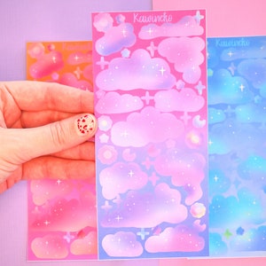 May include: Three sheets of iridescent stickers featuring clouds, stars, and other celestial designs. The sheets are pink, purple, and blue, with a gradient effect. The text "Kawinecko" appears on each sheet.