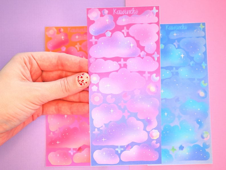May include: Three sheets of iridescent stickers featuring clouds, stars, and other celestial designs. The sheets are pink, purple, and blue, with a gradient effect. The text "Kawinecko" appears on each sheet.