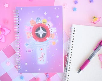 May include: Spiral-bound notebook with a purple cover featuring a cartoon design of a light stick with the word "Stay". The notebook is next to a blank notebook and a pink pen on a pink surface.