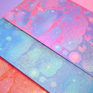 May include: Three sheets of iridescent paper with a galaxy theme. The paper is pink, blue, and orange with glitter and clouds. The word "Kawaii" is written on the top left corner of the blue sheet.