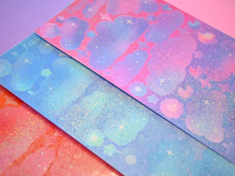 May include: Three sheets of iridescent paper with a galaxy theme. The paper is pink, blue, and orange with glitter and clouds. The word "Kawaii" is written on the top left corner of the blue sheet.
