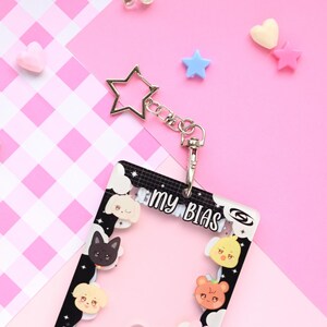 May include: A clear plastic ID badge holder with a black and white border, featuring cartoon animal faces and the text "my BIAS." It includes a silver star-shaped keychain and a clasp. The background is pink with scattered heart and star-shaped decorations.