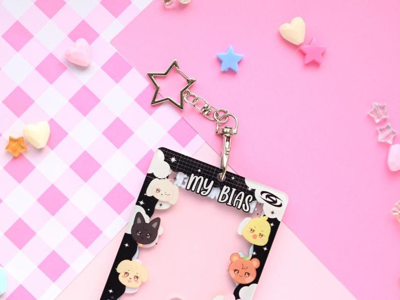 May include: A clear plastic ID badge holder with a black and white border, featuring cartoon animal faces and the text "my BIAS." It includes a silver star-shaped keychain and a clasp. The background is pink with scattered heart and star-shaped decorations.