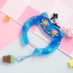 May include: A blue acrylic carabiner clip with a cartoon cat design, orange eyes, and a witch hat. The clip has a silver clasp and a small charm with a plant design. Decorative stars and a chain are also present.