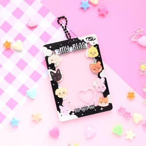 May include: A clear plastic card holder with a black border featuring cartoon animal faces and the text "MY BIAS". The holder has a chain and is surrounded by colorful star and heart-shaped decorations on a pink background.