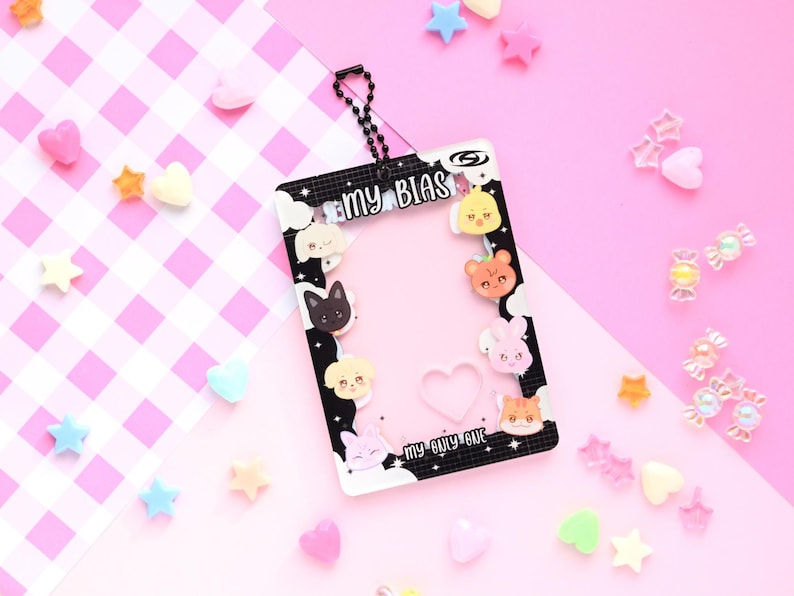 May include: A clear plastic card holder with a black border featuring cartoon animal faces and the text "MY BIAS". The holder has a chain and is surrounded by colorful star and heart-shaped decorations on a pink background.