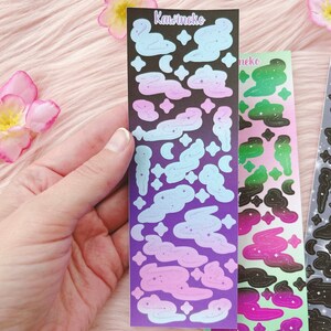 K&ouml;nnte beinhalten: Drei Bl&auml;tter mit Aufklebern zum Thema Galaxie. Die Aufkleber sind schwarz, lila, gr&uuml;n und pink mit wei&szlig;en Sternen und Wolken. Die Aufkleber sind mit "Kawineko" beschriftet.