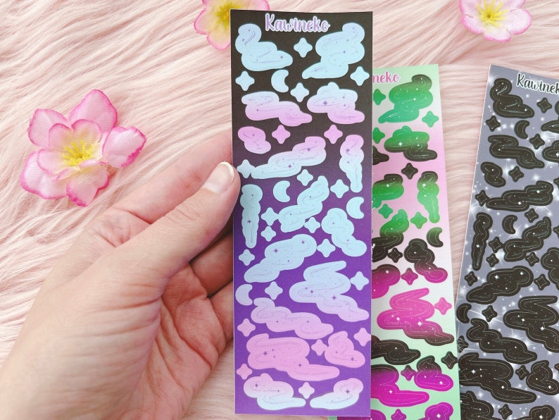K&ouml;nnte beinhalten: Drei Bl&auml;tter mit Aufklebern zum Thema Galaxie. Die Aufkleber sind schwarz, lila, gr&uuml;n und pink mit wei&szlig;en Sternen und Wolken. Die Aufkleber sind mit "Kawineko" beschriftet.