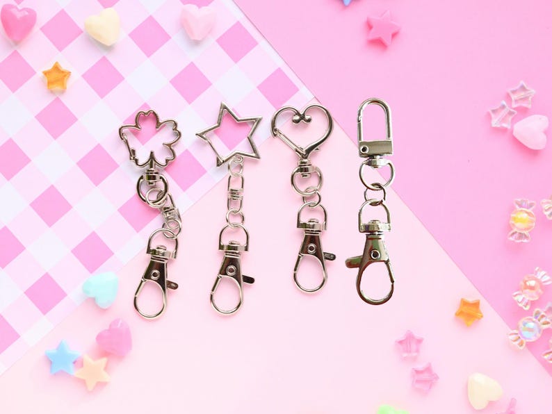 May include: Four silver-toned keychains with different shapes: a flower, a star, a heart, and a simple clasp. Each keychain has a lobster clasp for easy attachment. The background is a pink and white checkered pattern with scattered decorative elements.
