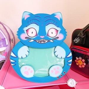 May include: A blue cat-shaped backpack with a large clear window on the belly, pink-rimmed eyes, sharp teeth, and light blue paws. The design features dark blue stripes on the sides and a smooth, rounded silhouette with three metal grommets.