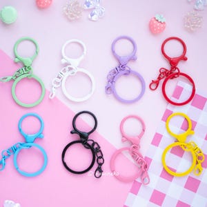 May include: A collection of colorful keychains in various shades including green, white, purple, red, blue, black, pink, and yellow. Each keychain features a round ring and a clasp. The keychains are arranged on a pink surface with decorative elements.