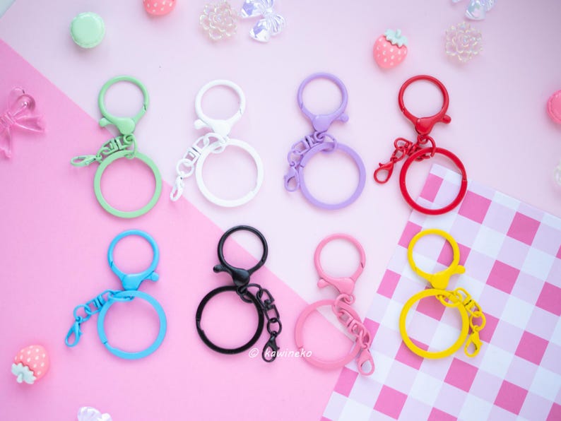 May include: A collection of colorful keychains in various shades including green, white, purple, red, blue, black, pink, and yellow. Each keychain features a round ring and a clasp. The keychains are arranged on a pink surface with decorative elements.