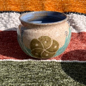 May include: A small, round, ceramic pot with a speckled glaze in shades of brown, white, and blue. The pot features a green and white floral design.
