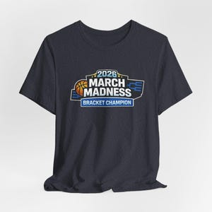 May include: Dark gray t-shirt featuring a basketball-themed graphic. The graphic includes the text "2026 March Madness Bracket Champion" in blue, white, and orange. The shirt has short sleeves and a crew neck.
