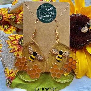 May include: Clear, teardrop-shaped earrings with gold glitter, featuring a black and yellow bee with rhinestone wings and a honeycomb design. The earrings are approximately 2 inches long and 1.5 inches wide.