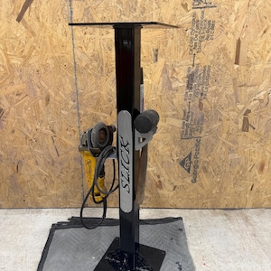 May include: A black metal stand with a square base and top, featuring a vertical support with the word "SLICK" in white. A yellow and black grinder and other tools are attached to the stand. The stand is on a gray surface.