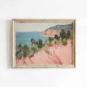 May include: A framed landscape painting depicting a coastal scene with a pink foreground, green trees, a blue ocean, and distant cliffs under a pale sky. The artwork is enclosed in a light-coloured wooden frame with a visible grain texture.