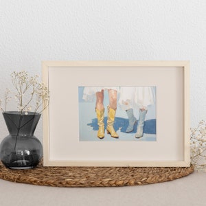 May include: A framed painting of two pairs of legs wearing boots. One pair is wearing yellow cowboy boots and the other pair is wearing blue boots. The painting is on a light blue background.