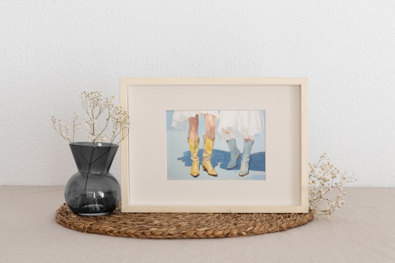 May include: A framed painting of two pairs of legs wearing boots. One pair is wearing yellow cowboy boots and the other pair is wearing blue boots. The painting is on a light blue background.