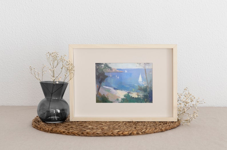 May include: A framed painting of a seascape with a sailboat in the distance. The painting is in a light wood frame and is set on a woven mat.