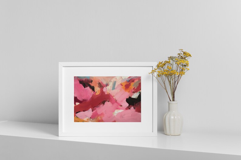 May include: Abstract art print in a white frame. The artwork features a vibrant mix of pink, red, and black brushstrokes. A small white vase with yellow flowers is placed to the right of the framed art, creating a balanced composition.