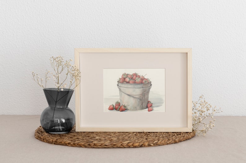 May include: A framed watercolor painting of a metal bucket filled with red strawberries. The bucket is sitting on a white surface. The painting is framed in a light wood frame.