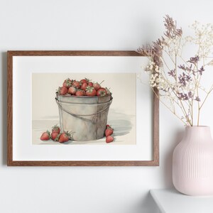 May include: A framed watercolor painting of a metal bucket filled with red strawberries. The bucket is sitting on a white surface with a few strawberries scattered around it.