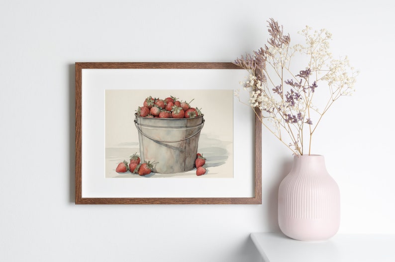 May include: A framed watercolor painting of a metal bucket filled with red strawberries. The bucket is sitting on a white surface with a few strawberries scattered around it.