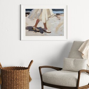 May include: An abstract art print in a white frame depicts a person walking on a beach. The painting features a white dress and white shoes with gold accents. The background shows the ocean and sand, with a palette of whites, blues, and tans.