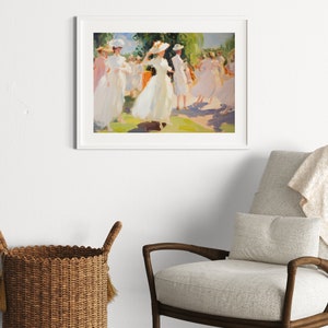 May include: A framed art print depicts a sunny outdoor scene of people in white dresses and hats. The impressionistic painting has a soft color palette of white, pink, green, and yellow. The artwork is displayed on a white wall above a chair and basket.