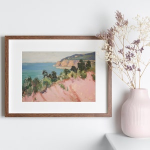 May include: A framed landscape painting depicting a coastal scene with pink cliffs, green trees, and a blue ocean under a pale sky. The artwork is housed in a rectangular wooden frame with a white mat. A light pink vase with dried flowers sits nearby.