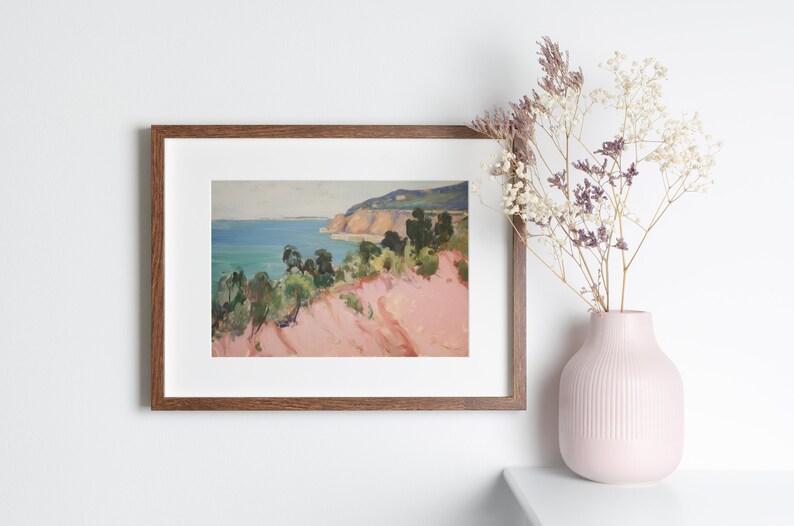 May include: A framed landscape painting depicting a coastal scene with pink cliffs, green trees, and a blue ocean under a pale sky. The artwork is housed in a rectangular wooden frame with a white mat. A light pink vase with dried flowers sits nearby.