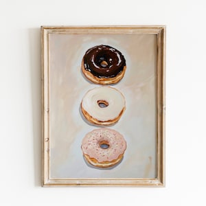 Op de afbeelding: Een ingelijst schilderij van drie donuts die verticaal op elkaar gestapeld zijn. De bovenste donut is geglazuurd met chocolade glazuur, de middelste donut is geglazuurd met wit glazuur en de onderste donut is geglazuurd met roze glazuur en sprinkles.