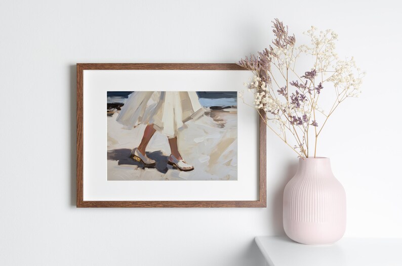 May include: A framed art print depicts a person walking on a beach, wearing a white dress and white shoes. The artwork is in a brown frame and is displayed on a white wall. A pink vase with dried flowers sits on a white surface.
