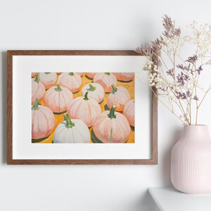 Op de afbeelding: Een ingelijste kunstdruk met een schilderij van meerdere lichtroze en witte pompoenen met groene stelen op een felgele achtergrond, weergegeven in een donkerhouten lijst naast een lichtroze vaas met gedroogde bloemen.