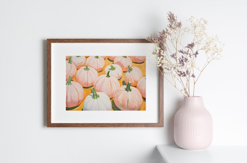 Op de afbeelding: Een ingelijste kunstdruk met een schilderij van meerdere lichtroze en witte pompoenen met groene stelen op een felgele achtergrond, weergegeven in een donkerhouten lijst naast een lichtroze vaas met gedroogde bloemen.