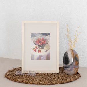 May include: A framed painting of a bowl of red strawberries on a white tablecloth. The bowl is on a table with a fork and a few strawberries scattered around it. The painting is in a white frame and is hanging on a white wall.