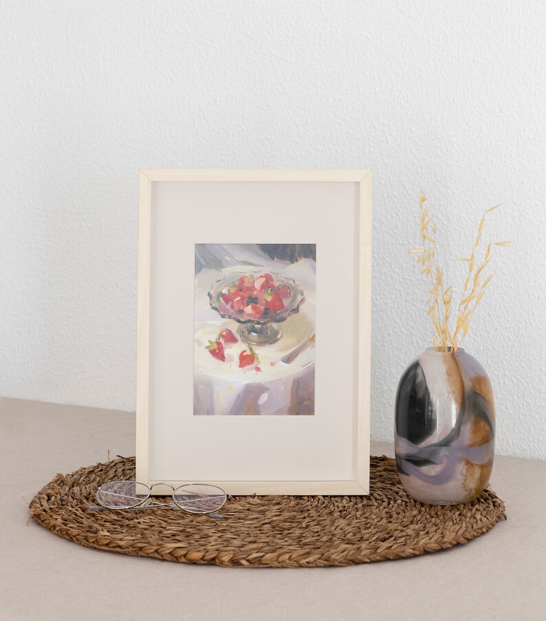 May include: A framed painting of a bowl of red strawberries on a white tablecloth. The bowl is on a table with a fork and a few strawberries scattered around it. The painting is in a white frame and is hanging on a white wall.