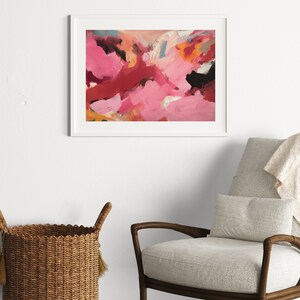 May include: Abstract art print in a white frame. The artwork features a vibrant mix of pink, red, orange, and black brushstrokes. The framed art is displayed above a gray armchair with a beige throw blanket and a woven basket.