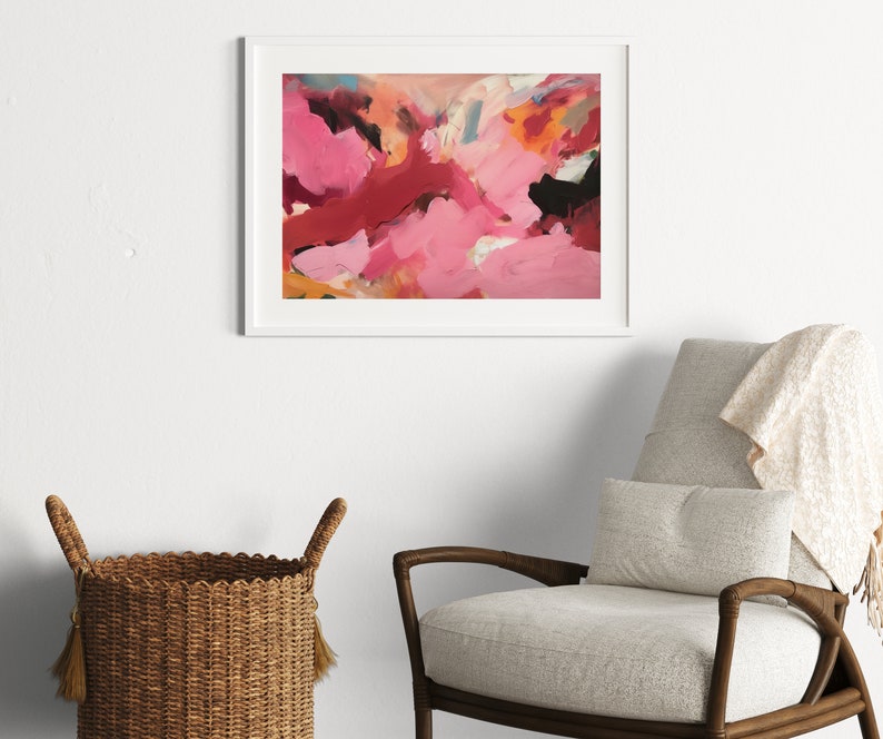 May include: Abstract art print in a white frame. The artwork features a vibrant mix of pink, red, orange, and black brushstrokes. The framed art is displayed above a gray armchair with a beige throw blanket and a woven basket.
