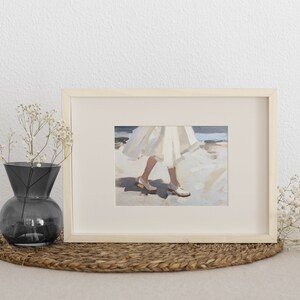 May include: Framed art print featuring a painting of a person's legs and feet in white shoes and a flowing white dress. The artwork is displayed in a light wood frame. A dark gray vase with white flowers sits to the left.