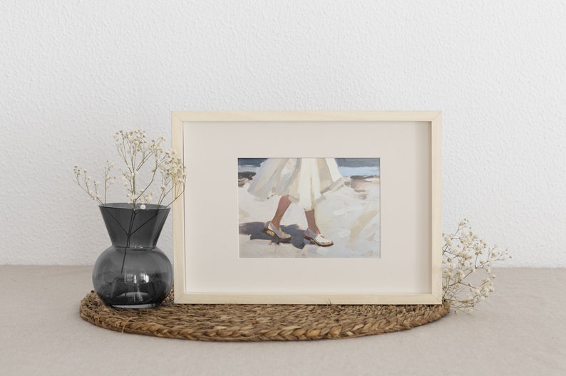 May include: Framed art print featuring a painting of a person's legs and feet in white shoes and a flowing white dress. The artwork is displayed in a light wood frame. A dark gray vase with white flowers sits to the left.