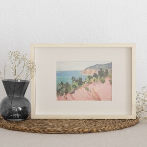 May include: A framed landscape painting featuring a coastal scene with pink cliffs, green trees, and a blue sea. The artwork is housed in a light-coloured wooden frame with a cream-coloured mat. A dark grey glass vase with white flowers sits nearby.