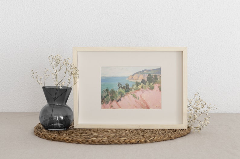 May include: A framed landscape painting featuring a coastal scene with pink cliffs, green trees, and a blue sea. The artwork is housed in a light-coloured wooden frame with a cream-coloured mat. A dark grey glass vase with white flowers sits nearby.