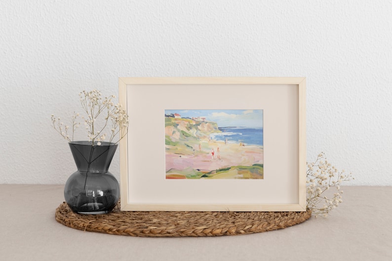 Puede incluir: Una pintura enmarcada que representa una escena de playa con un acantilado, el oc&eacute;ano y personas en la arena. La pintura est&aacute; en un marco de madera clara y tiene un passe-partout blanco.