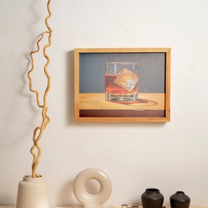 May include: A framed illustration of a glass of whiskey with ice on a wooden surface. The frame is made of light wood and has a white mat. The illustration is set against a blue background. The image is displayed on a white wall with a vase of dried branches and a ceramic sculpture on a table.