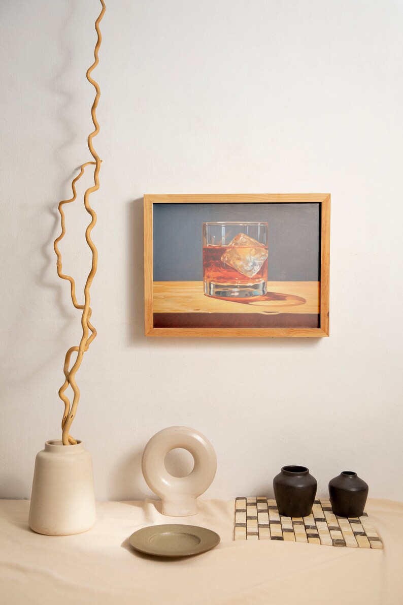 May include: A framed illustration of a glass of whiskey with ice on a wooden surface. The frame is made of light wood and has a white mat. The illustration is set against a blue background. The image is displayed on a white wall with a vase of dried branches and a ceramic sculpture on a table.