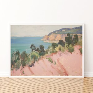 May include: A framed landscape painting featuring a coastal scene with a pink foreground, green trees, and a blue sea meeting a distant cliffside under a pale sky. The artwork is enclosed in a simple white frame, displaying soft brushstrokes.
